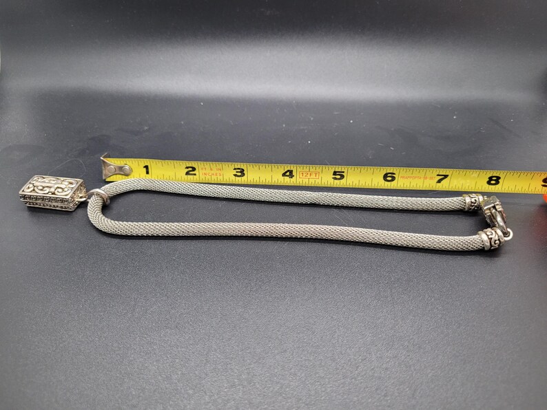 May include: A silver-toned mesh chain necklace featuring a rectangular pendant with an intricate scrollwork pattern. A yellow measuring tape is positioned alongside the necklace, indicating a total length of approximately 8 inches for the chain.