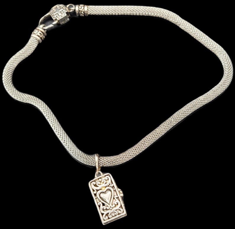 May include: A silver-toned mesh chain necklace featuring a rectangular pendant with an embossed heart design and intricate scrollwork. The necklace includes a decorative clasp with a cross motif, showcasing a metallic finish and a flexible link structure.