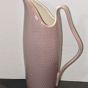 May include: A tall, light purple ceramic pitcher with a white interior and a white handle. The pitcher has a textured, ribbed design.