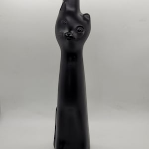 May include: A matte black glass bottle shaped like a stylized cat. The bottle features a tall, slender body that forms the cat's torso, with a molded head at the top including ears, eyes, and whiskers. The neck of the bottle extends from the cat's head.