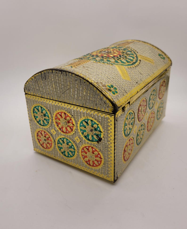 Puede incluir: Un cofre decorativo de metal con tapa abovedada. El cofre es plateado con ribetes dorados y presenta dise&ntilde;os florales coloridos en rojo, verde y amarillo. El cofre tiene una base rectangular y una parte superior curva.