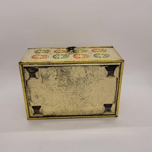 Puede incluir: Una caja met&aacute;lica rectangular vintage con tapa abisagrada. La caja es dorada con un patr&oacute;n decorativo de c&iacute;rculos rojos, verdes y amarillos en la tapa. La caja tiene un cierre de pestillo y cuatro patas peque&ntilde;as.