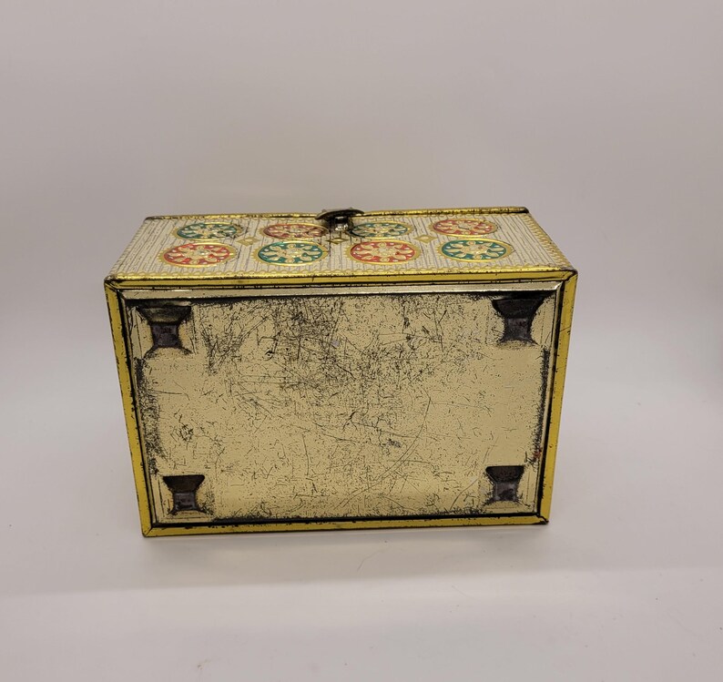 Puede incluir: Una caja met&aacute;lica rectangular vintage con tapa abisagrada. La caja es dorada con un patr&oacute;n decorativo de c&iacute;rculos rojos, verdes y amarillos en la tapa. La caja tiene un cierre de pestillo y cuatro patas peque&ntilde;as.