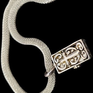 May include: A silver-toned snake chain necklace featuring a rectangular pendant with an intricate, raised scrollwork design that incorporates a central cross motif. The chain has a fine, woven texture and is connected to the metal pendant by a small loop.