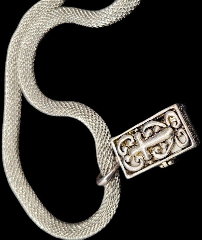 May include: A silver-toned snake chain necklace featuring a rectangular pendant with an intricate, raised scrollwork design that incorporates a central cross motif. The chain has a fine, woven texture and is connected to the metal pendant by a small loop.