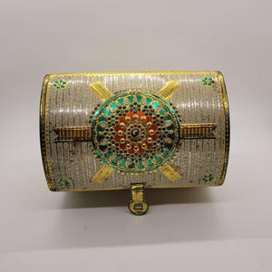 Puede incluir: Un cofre decorativo plateado con ribetes dorados y un dise&ntilde;o circular central en rojo, verde azulado y dorado. El cofre tiene forma rectangular con la parte superior curvada y un peque&ntilde;o cierre de pestillo. Se ven detalles florales.