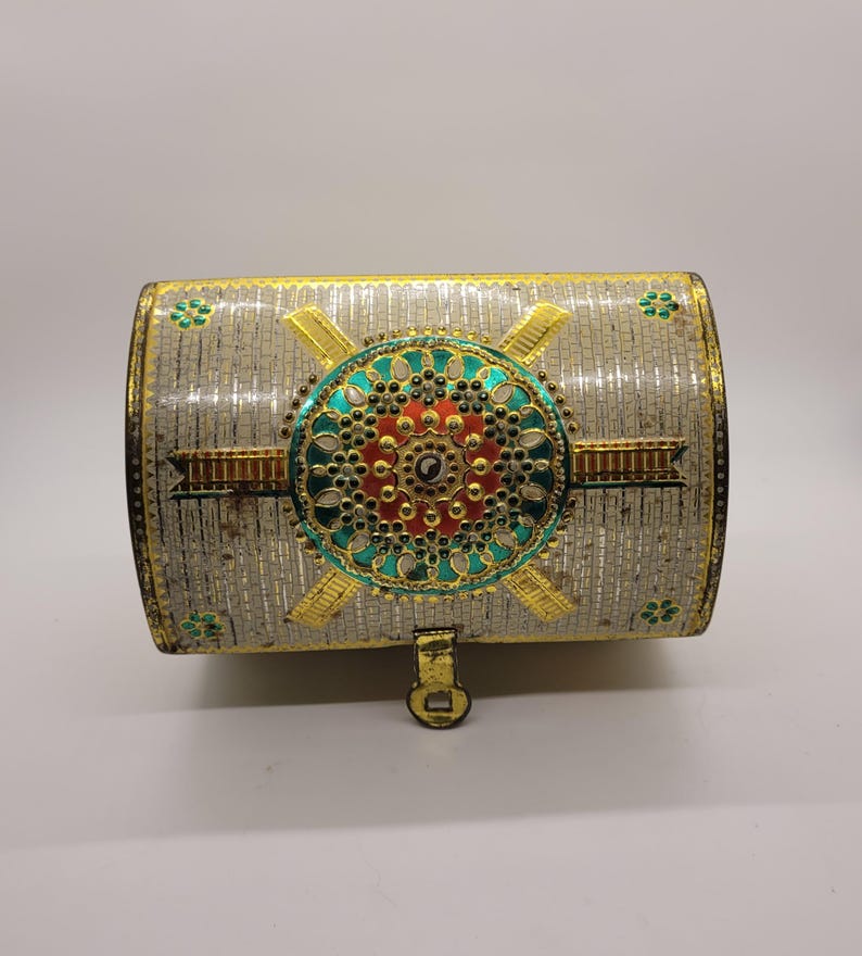 Puede incluir: Un cofre decorativo plateado con ribetes dorados y un dise&ntilde;o circular central en rojo, verde azulado y dorado. El cofre tiene forma rectangular con la parte superior curvada y un peque&ntilde;o cierre de pestillo. Se ven detalles florales.