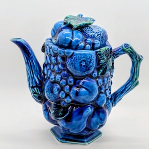 May include: A cobalt blue ceramic teapot with a lid, handle, and spout. The body is decorated with a textured fruit design, including grapes and other fruits. The handle and spout have a branch-like appearance. The lid is topped with a green leaf.