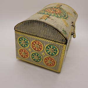 Puede incluir: Un cofre decorativo de metal con tapa curvada y base rectangular. El cofre es plateado con ribetes dorados y presenta dise&ntilde;os florales coloridos en rojo, verde y dorado. Un peque&ntilde;o cierre asegura la tapa.