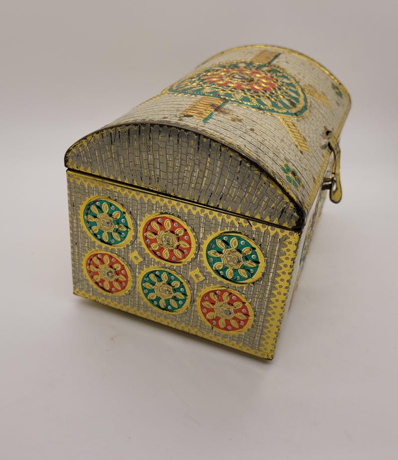 Puede incluir: Un cofre decorativo de metal con tapa curvada y base rectangular. El cofre es plateado con ribetes dorados y presenta dise&ntilde;os florales coloridos en rojo, verde y dorado. Un peque&ntilde;o cierre asegura la tapa.