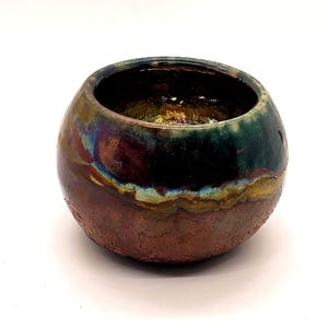 May include: A small, round ceramic bowl with a textured, earthy brown base and a dark green rim. The bowl's surface has iridescent hues of gold, purple, and blue. The interior is dark, suggesting a glazed finish. This decorative bowl is suitable for home decor.