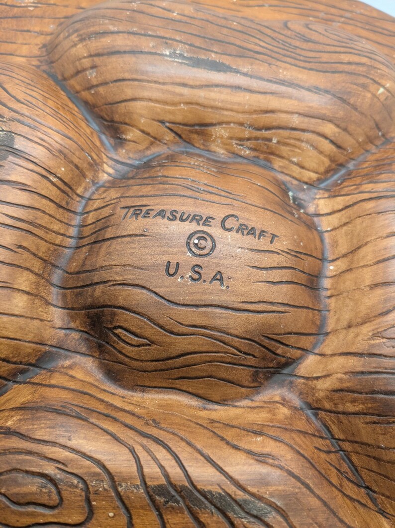 May include: A brown, wooden-textured serving dish with multiple compartments. The dish is marked with the text "TREASURE CRAFT" and "U.S.A.". The dish has a rustic, natural appearance, suitable for serving snacks or appetizers.