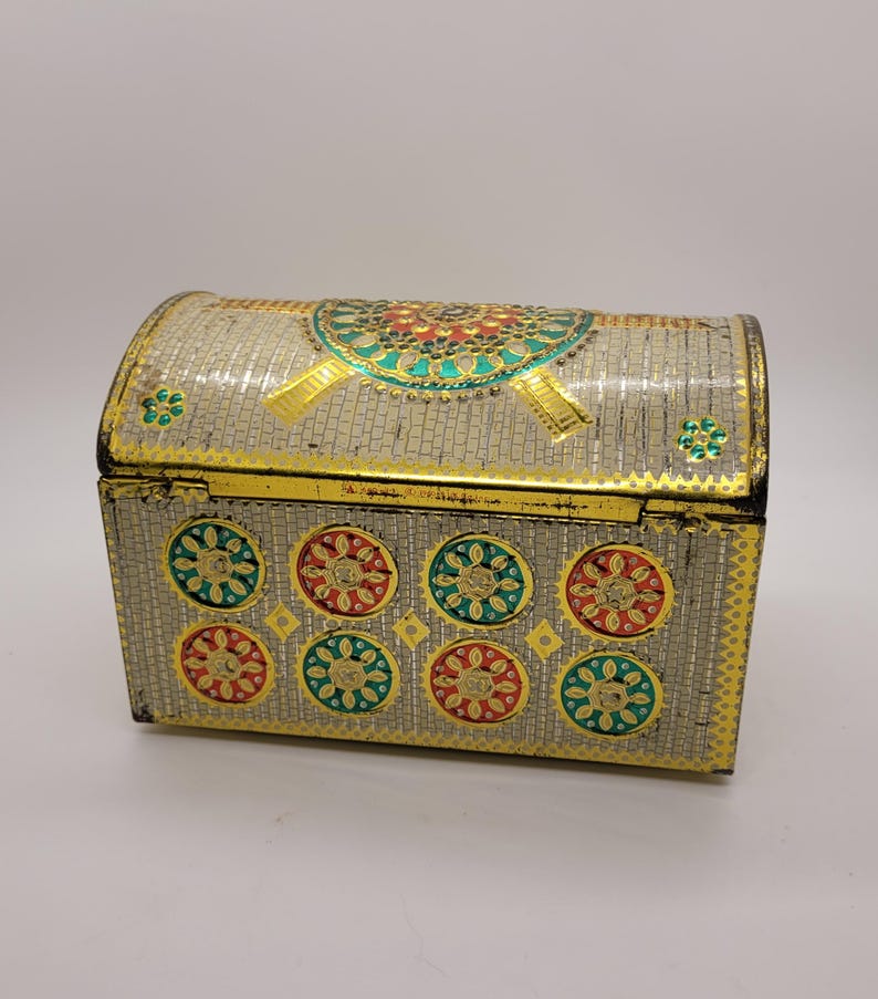 Puede incluir: Un cofre decorativo de metal con tapa abovedada. El cofre es plateado con adornos dorados y presenta dise&ntilde;os florales coloridos en rojo, verde y dorado. El cofre probablemente se utiliza para guardar objetos peque&ntilde;os.