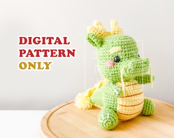 May include: A crocheted green and yellow dragon amigurumi toy with a cute face. The dragon is sitting on a wooden base. DIGITAL PATTERN ONLY.