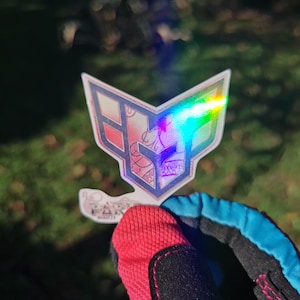 Może przedstawiać: Holograficzna naklejka z różowo-białym wzorem przedstawiającym stylizowaną literę "F" z geometrycznym wzorem. Naklejka ma tęczowy, opalizujący wygląd.