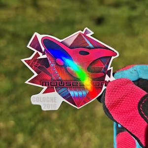May include: A holographic sticker with a geometric design featuring a stylized bird head in pink and red, with a rainbow gradient. The sticker has the text "mouses art" and "COLOGNE 2016".