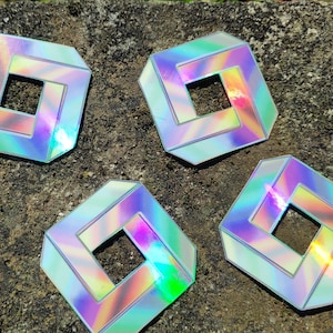 Puede incluir: Cuatro pegatinas holográficas iridiscentes con un diseño cuadrado. Cada pegatina presenta un cuadrado con un cuadrado más pequeño en el interior, creando una ilusión óptica de un cubo.