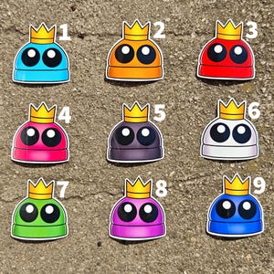 May include: Nine colorful stickers featuring a cartoon character with a crown. Each sticker has a different color scheme, including blue, orange, red, pink, purple, white, green, and a darker blue. The character has large black eyes and a yellow crown.