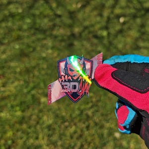 K&ouml;nnte beinhalten: Ein farbenfroher Aufkleber mit einem teufelartigen Charakter mit Kopfh&ouml;rern, einem Laserstrahl und dem Text "3D ZETA". Der Aufkleber wird von einem rot-schwarzen Handschuh mit blauen Akzenten vor einem gr&uuml;nen, grasbewachsenen Hintergrund gehalten.