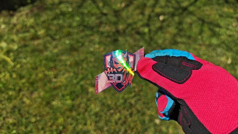 K&ouml;nnte beinhalten: Ein farbenfroher Aufkleber mit einem teufelartigen Charakter mit Kopfh&ouml;rern, einem Laserstrahl und dem Text "3D ZETA". Der Aufkleber wird von einem rot-schwarzen Handschuh mit blauen Akzenten vor einem gr&uuml;nen, grasbewachsenen Hintergrund gehalten.