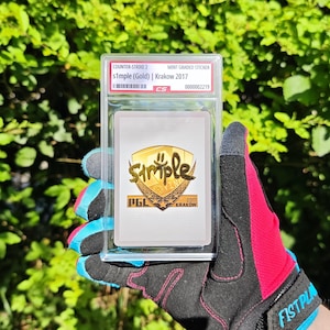 May include: A mint graded sticker for Counter-Strike 2, featuring s1mple (Gold) from Krakow 2017. The sticker is encased in a clear protective holder. The background is a blurred view of green foliage. The holder is being held by a person wearing a black, pink, and blue glove.
