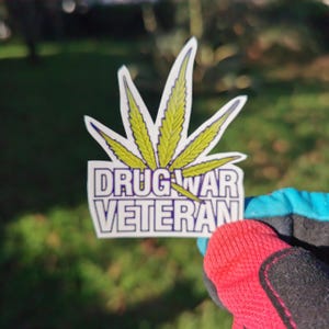 以下が含まれることがあります： 緑色のマリファナの葉と「Drug War Veteran」という青い文字と白い文字が書かれた白いステッカーです