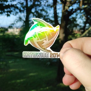 May include: A holographic sticker with a green, blue, and gold design. The sticker features a stylized globe with a three-dimensional effect. The text "KATOWICE 2014" is printed below the globe.