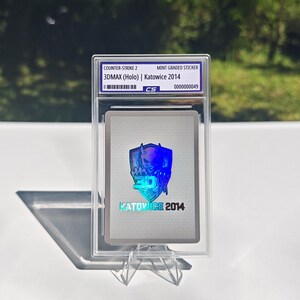 Pu&ograve; includere: Un adesivo di Counter-Strike 2, valutato come nuovo, con un design olografico 3DMAX di Katowice 2014. L'adesivo &egrave; racchiuso in un supporto protettivo trasparente, esposto su un supporto trasparente. Il testo "CS" &egrave; visibile.