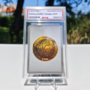 Puede incluir: Una pegatina de grado menta para Counter-Strike 2, con "DickStacy (Gold) | Katowice 2019". La pegatina tiene un emblema dorado con el texto "DICK" y el logotipo "ESL". La pegatina est&aacute; dentro de un soporte de pl&aacute;stico transparente.