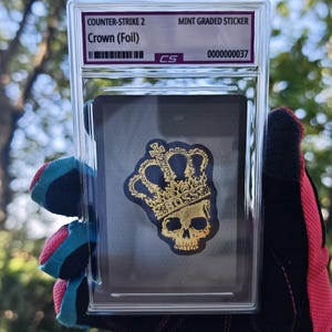 May include: A Counter-Strike 2 Crown (Foil) mint graded sticker encased in a clear plastic slab. The sticker features a gold-coloured skull wearing a crown with the word BOSS printed on the band, set against a black background with a barcode.