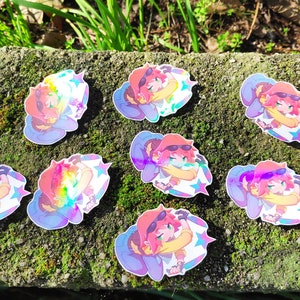 May include: Eight iridescent holographic stickers featuring a cartoon character with pink hair, wearing a blue and white outfit and a red hat. The character is holding a star and has a rainbow background.