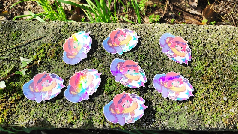 May include: Eight iridescent holographic stickers featuring a cartoon character with pink hair, wearing a blue and white outfit and a red hat. The character is holding a star and has a rainbow background.