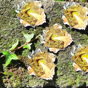 Op de afbeelding: Vijf gouden en gele draken stickers met een Keltisch knoopontwerp. De draak spuit vuur en de stickers bevinden zich op een mossig groen oppervlak.