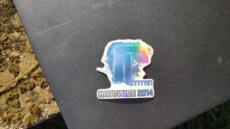 May include: A holographic sticker with a blue and white image of a door with the text "TITAN KATOWICE 2014" underneath.