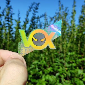Könnte beinhalten: Ein holografischer Aufkleber mit dem Wort "VOX" in grünen und gelben Buchstaben. Das "O" ist ein gelber Kreis mit einem schwarzen Gesicht mit einem Auge. Der Aufkleber ist diagonal und hat einen regenbogenfarbenen, schillernden Hintergrund.