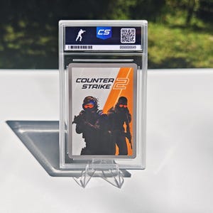 Pu&ograve; includere: Una carta collezionabile valutata con il logo e i personaggi del gioco "Counter-Strike 2". La carta &egrave; racchiusa in un supporto di plastica trasparente con un logo "CS" e un numero di serie. L'immagine mostra due figure in equipaggiamento tattico.