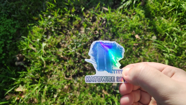 May include: A holographic sticker with a blue and white design. The sticker features a stylized image of a building with the text "KATOWICE 2019" below it.