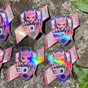 K&ouml;nnte beinhalten: Eine Sammlung von Aufklebern mit einem rosa Teufel-Charakter mit Kopfh&ouml;rern und dem Text "3D" auf einem Schild. Die Aufkleber haben einen holografischen Effekt und liegen auf einer Stein Oberfl&auml;che.