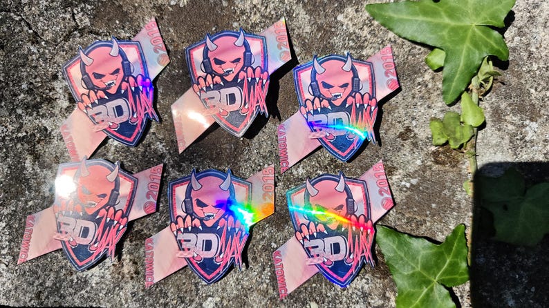 K&ouml;nnte beinhalten: Eine Sammlung von Aufklebern mit einem rosa Teufel-Charakter mit Kopfh&ouml;rern und dem Text "3D" auf einem Schild. Die Aufkleber haben einen holografischen Effekt und liegen auf einer Stein Oberfl&auml;che.