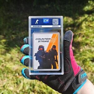 Pu&ograve; includere: Una carta collezionabile di Counter-Strike 2 in una custodia protettiva. La carta presenta due figure in equipaggiamento tattico con il logo del gioco. La custodia ha un'etichetta blu e bianca con un codice QR e un numero di serie.