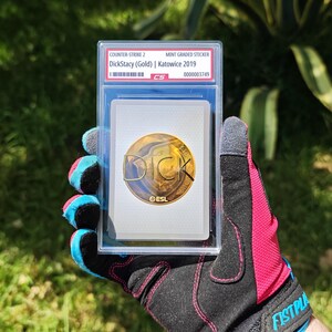 Puede incluir: Una pegatina de Counter-Strike 2 de grado superior con "DickStacy (Gold) | Katowice 2019". La pegatina est&aacute; dentro de un soporte protector transparente. La pegatina tiene un dise&ntilde;o dorado y blanco con el texto "DICK" y "ESL".
