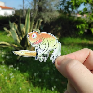 Op de afbeelding: Een cartoon kikker met een tandenrij houdt een mes in zijn hand. De kikker is geschilderd in tinten oranje, geel en groen. De kikker is een sticker.