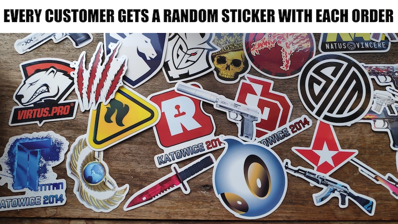 May include: A collection of colorful stickers featuring various logos and designs, including a red and white logo with the text "KATOWICE 2014", a black and white logo with the text "NATUS VINCERE", and a yellow and black warning sign with a flame design. The stickers are arranged on a wooden surface.