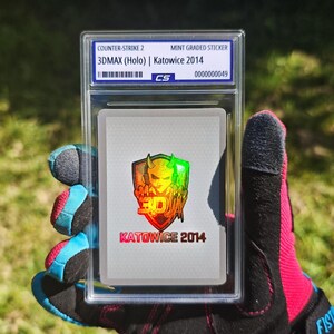 Pu&ograve; includere: Un adesivo di Counter-Strike 2 di grado menta, con un design olografico 3DMAX di Katowice 2014. L'adesivo &egrave; racchiuso in un supporto protettivo trasparente, con testo in alto e in basso. Il supporto &egrave; tenuto da una persona che indossa un guanto nero e rosa.