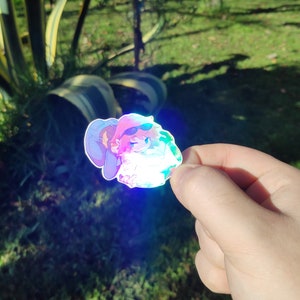 May include: A holographic sticker featuring a cartoon character wearing a hat and sunglasses. The character is holding a green object and has a pink and blue color scheme.