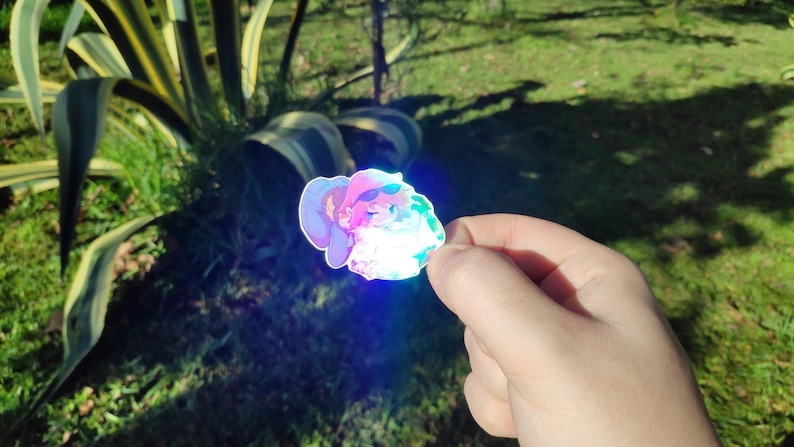 May include: A holographic sticker featuring a cartoon character wearing a hat and sunglasses. The character is holding a green object and has a pink and blue color scheme.