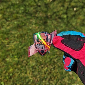K&ouml;nnte beinhalten: Ein farbenfroher Aufkleber mit einem Sch&auml;del mit Kopfh&ouml;rern, einem Schild und dem Text "3D". Der Aufkleber wird von einem rot-schwarzen Handschuh mit blauen Akzenten vor einem gr&uuml;nen Gras Hintergrund gehalten.