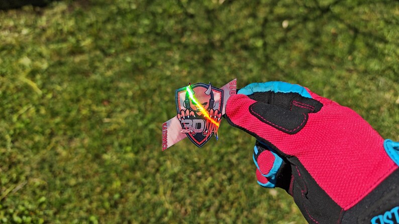 K&ouml;nnte beinhalten: Ein farbenfroher Aufkleber mit einem Sch&auml;del mit Kopfh&ouml;rern, einem Schild und dem Text "3D". Der Aufkleber wird von einem rot-schwarzen Handschuh mit blauen Akzenten vor einem gr&uuml;nen Gras Hintergrund gehalten.