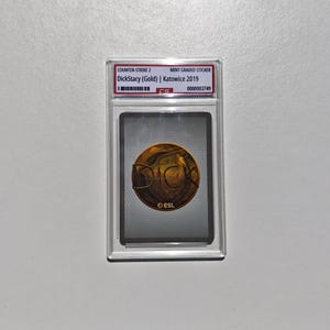 Puede incluir: Una pegatina Counter-Strike 2 DickStacy (Oro) Katowice 2019, calificada como nueva, en una funda protectora transparente. La pegatina presenta un emblema dorado con el texto "ESL". La funda tiene un n&uacute;mero de serie.