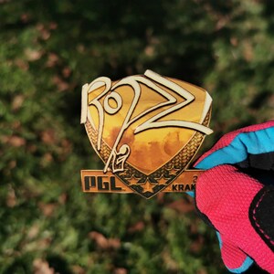 Könnte beinhalten: Eine goldfarbene Metallstifte mit einem schildförmigen Design. Der Schild hat eine weiße Inschrift mit der Aufschrift "RO22" und eine schwarze Inschrift mit der Aufschrift "PGL" und "KRAK".